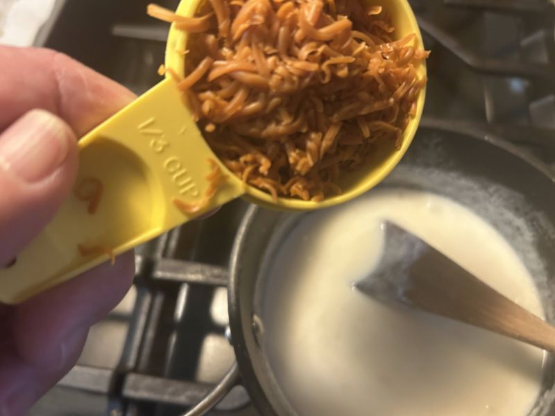 Grated Gjetost cheese added to creamy sauce in saucepan